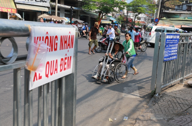 B&agrave; Nguyễn Thị Thiện (73 tuổi) đi xe lăn b&aacute;n v&eacute; số. Trước kia b&agrave; c&oacute; thể l&ecirc;n lề đi v&ograve;ng quanh vỉa h&egrave; b&aacute;n. Nhưng từ ng&agrave;y lắp r&agrave;o chắn, b&agrave; buộc phải b&aacute;n h&agrave;ng dưới l&ograve;ng đường.
