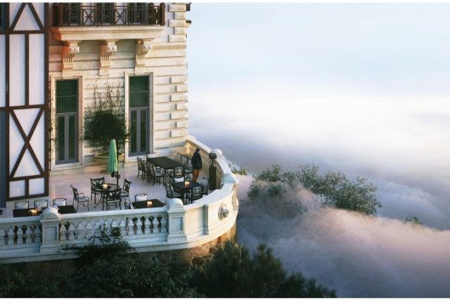 Tòa lâu đài nằm trên đỉnh núi, giữa không gian mây xanh bồng bềnh, cùng với những ngọn núi xanh xanh phía xa.
